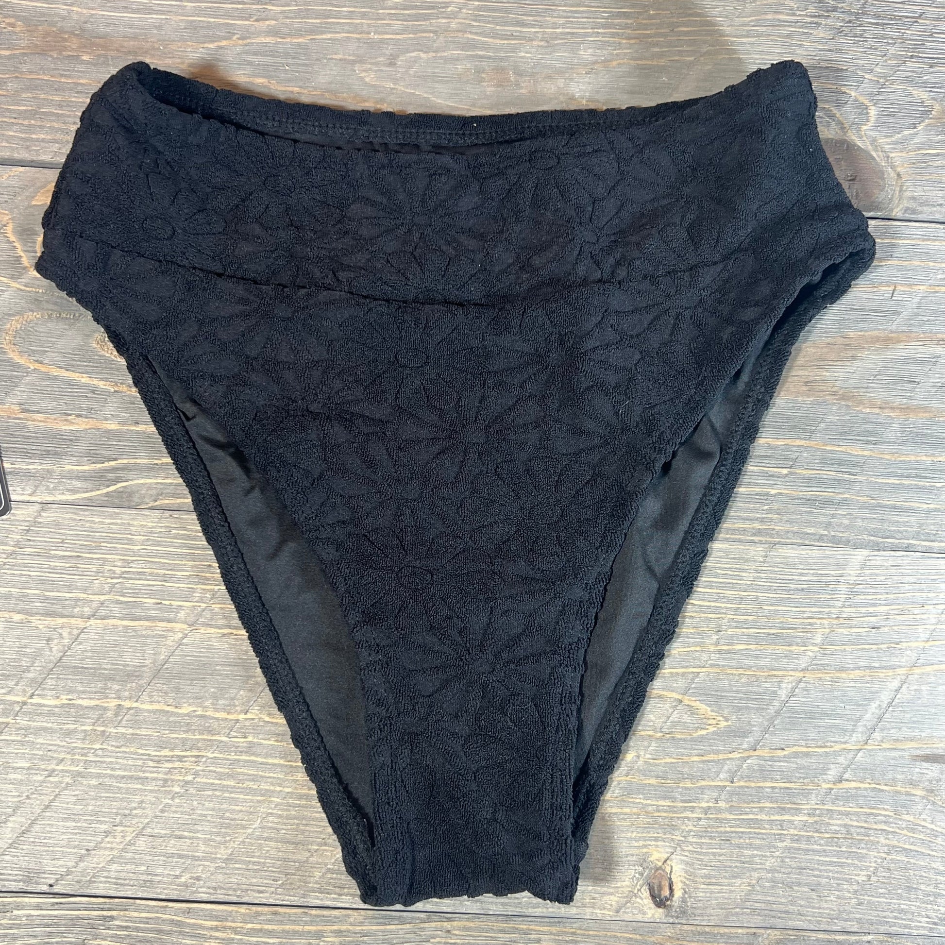 Black high-waisted bikini bottom with floral texture on a wooden background.