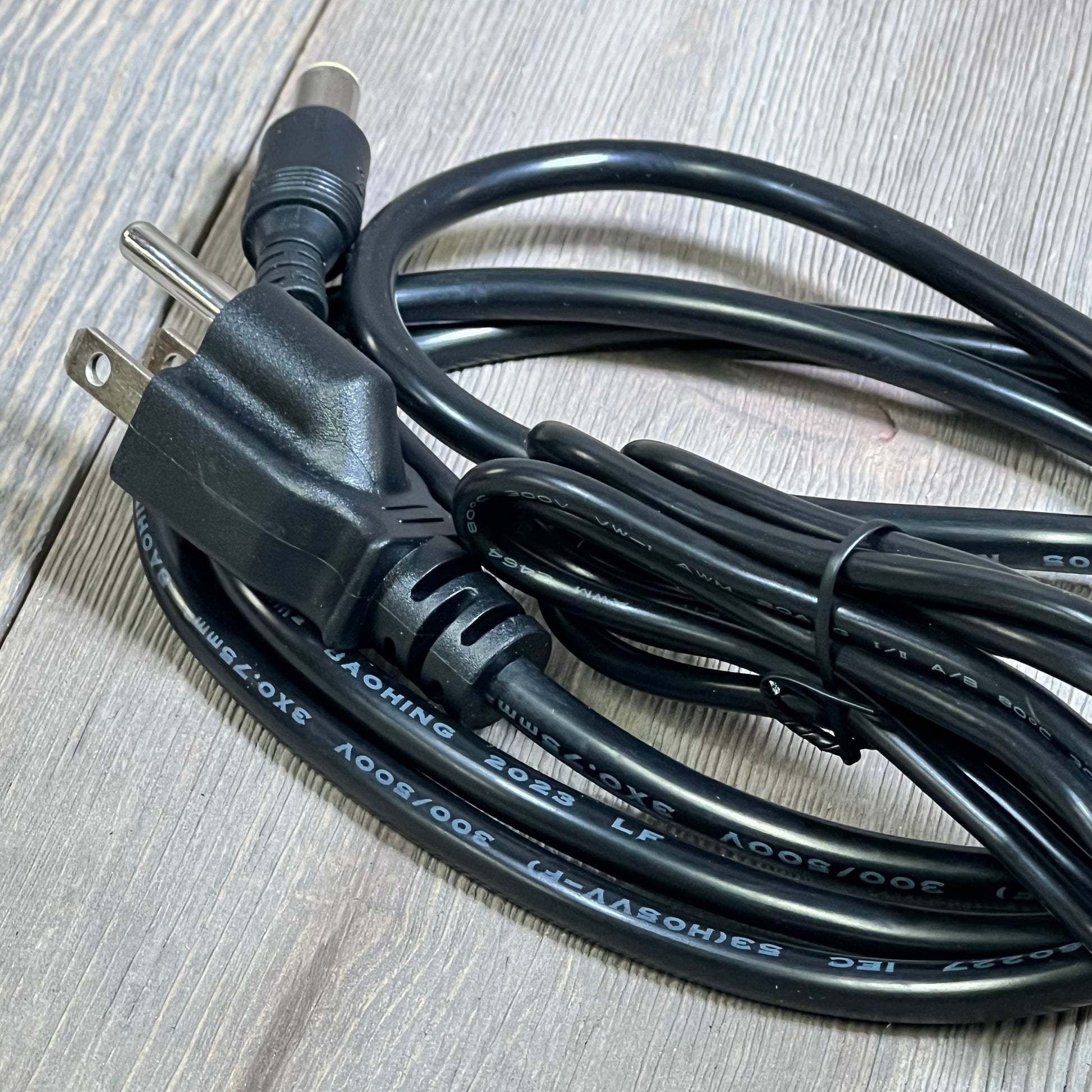 Black 24V 90W AC adapter cable for Jackery Portable Power Station, coiled on a wooden surface, showing dual plug design.