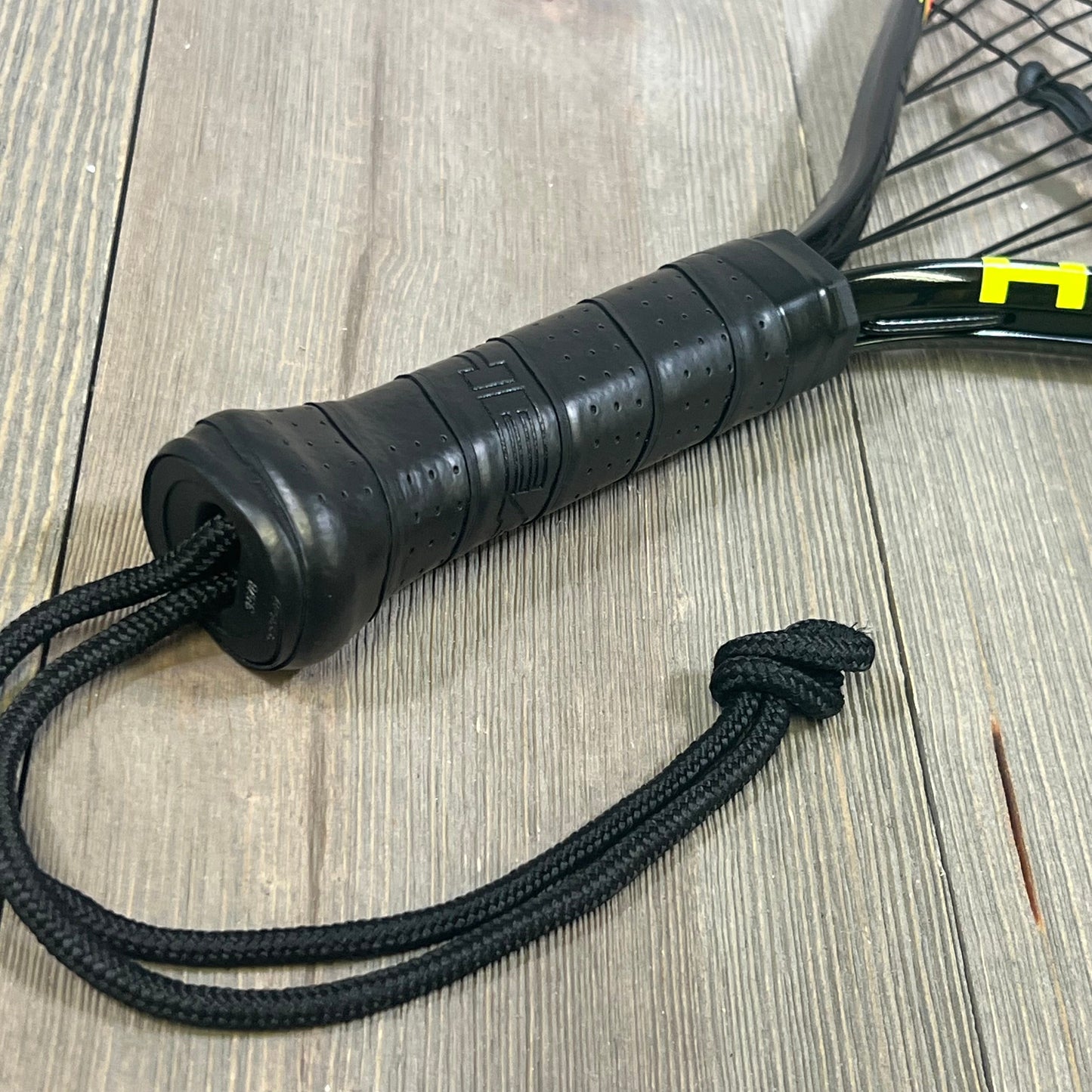 Close-up of 1047180423-HEAD CPS Heat Racquetball Racquet handle, featuring a black grip and wrist strap on wood surface.