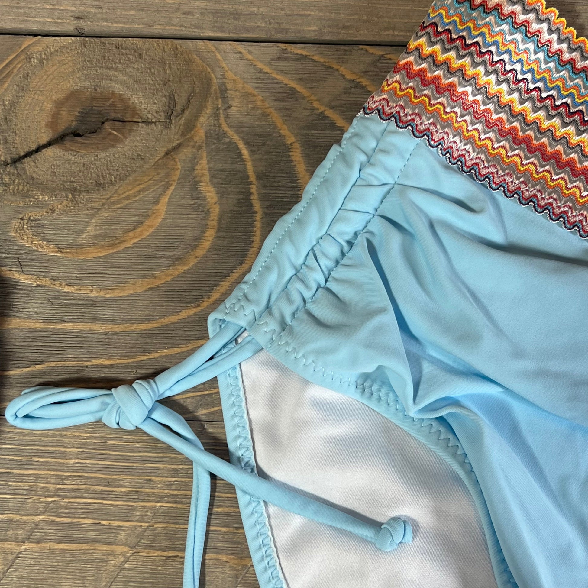 Blue high-waisted bikini bottom with colorful zigzag trim and side tie detail on wooden background.