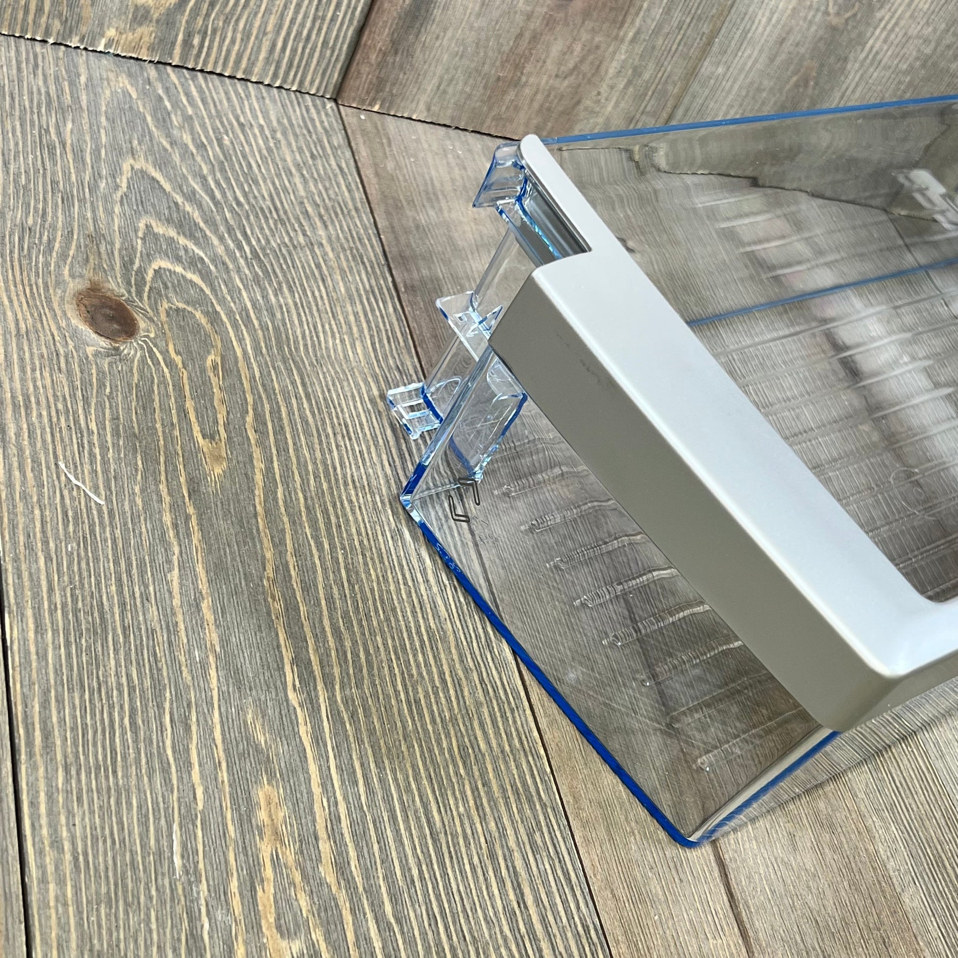 Clear plastic refrigerator door shelf bin compatible with Bosch 00673122, angled view on wooden surface.