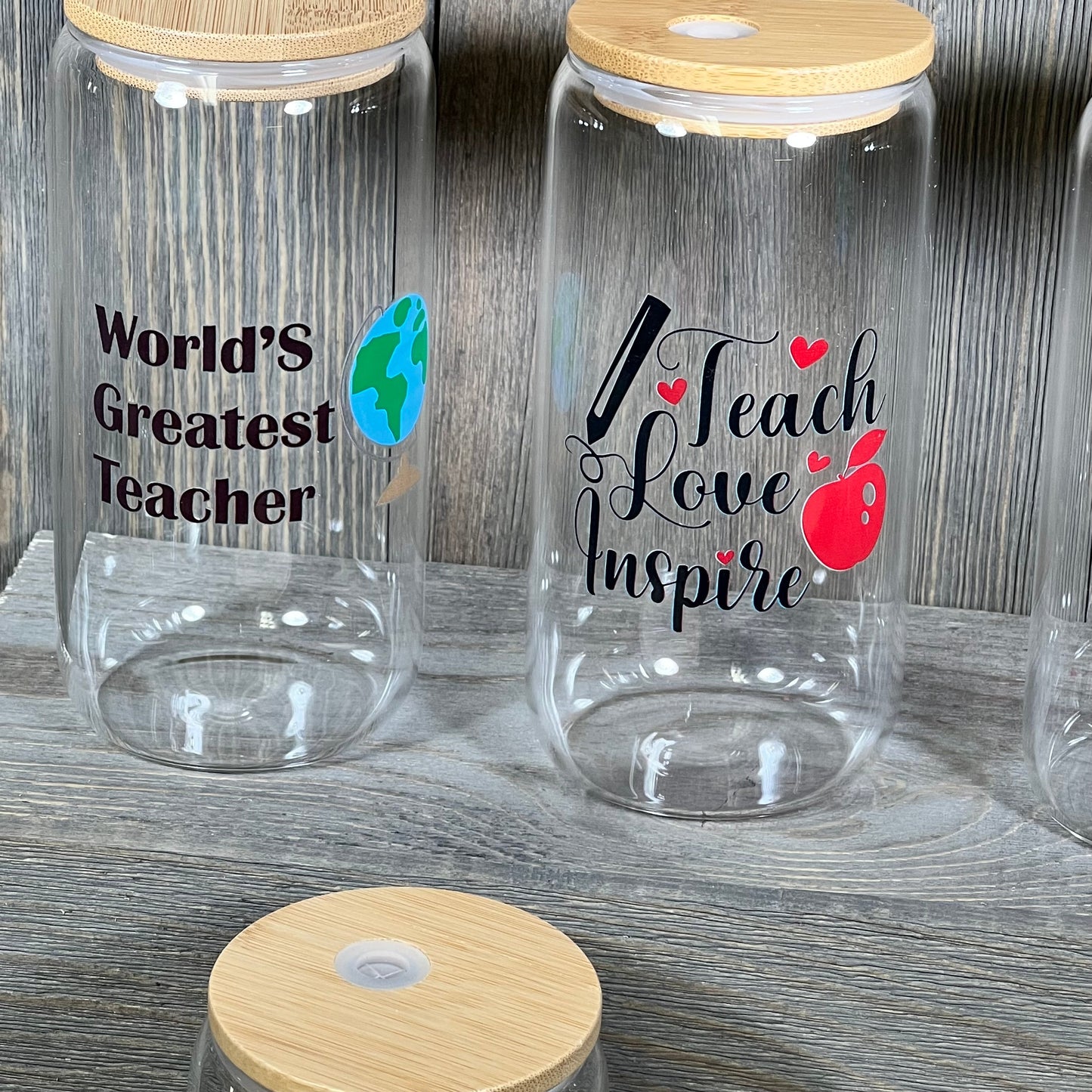 Glass cups with bamboo lids featuring "World's Greatest Teacher" and "Teach Love Inspire" designs, ideal as teacher gifts.