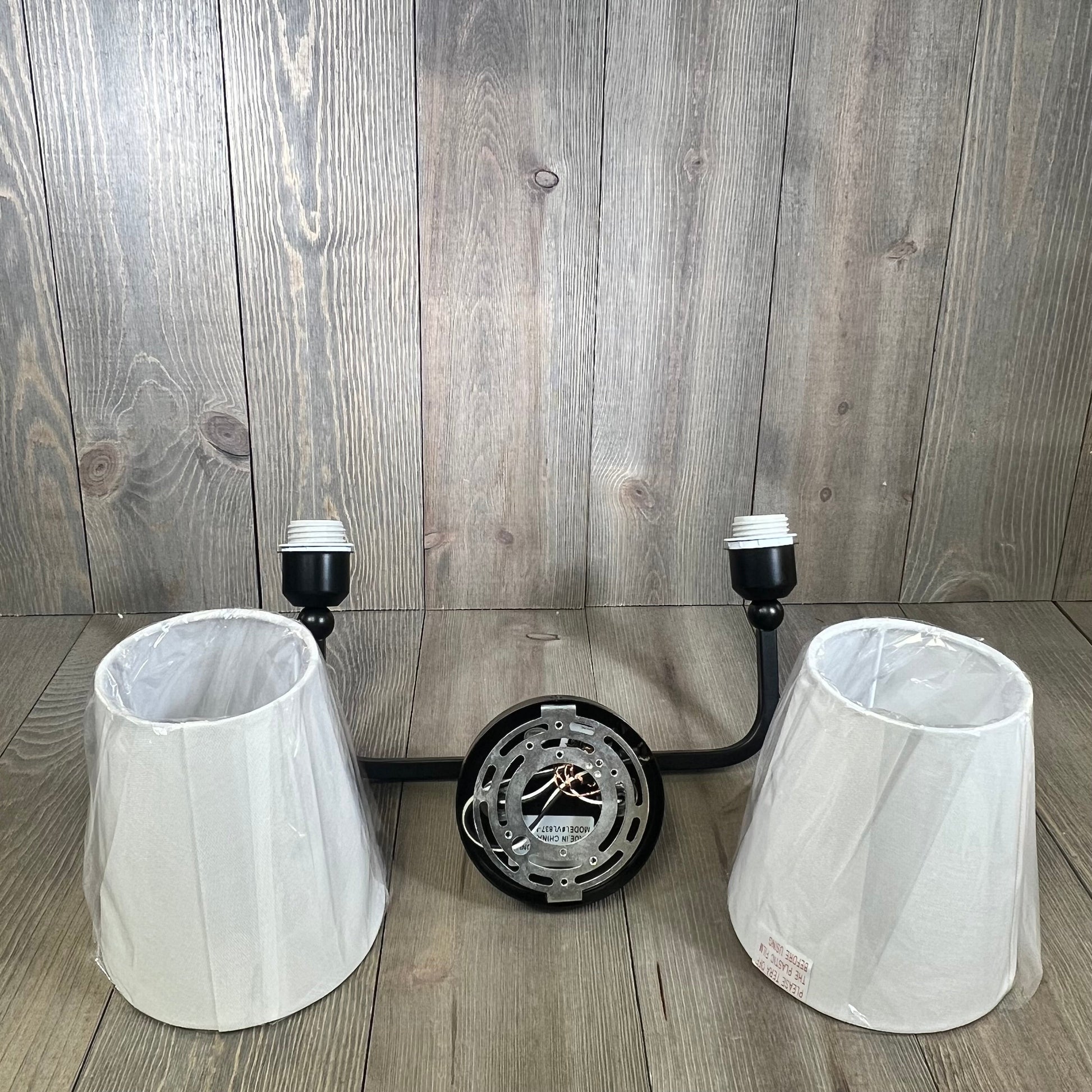 Matte black 2-light bathroom vanity fixture with white fabric shades, shown disassembled on a wooden surface.