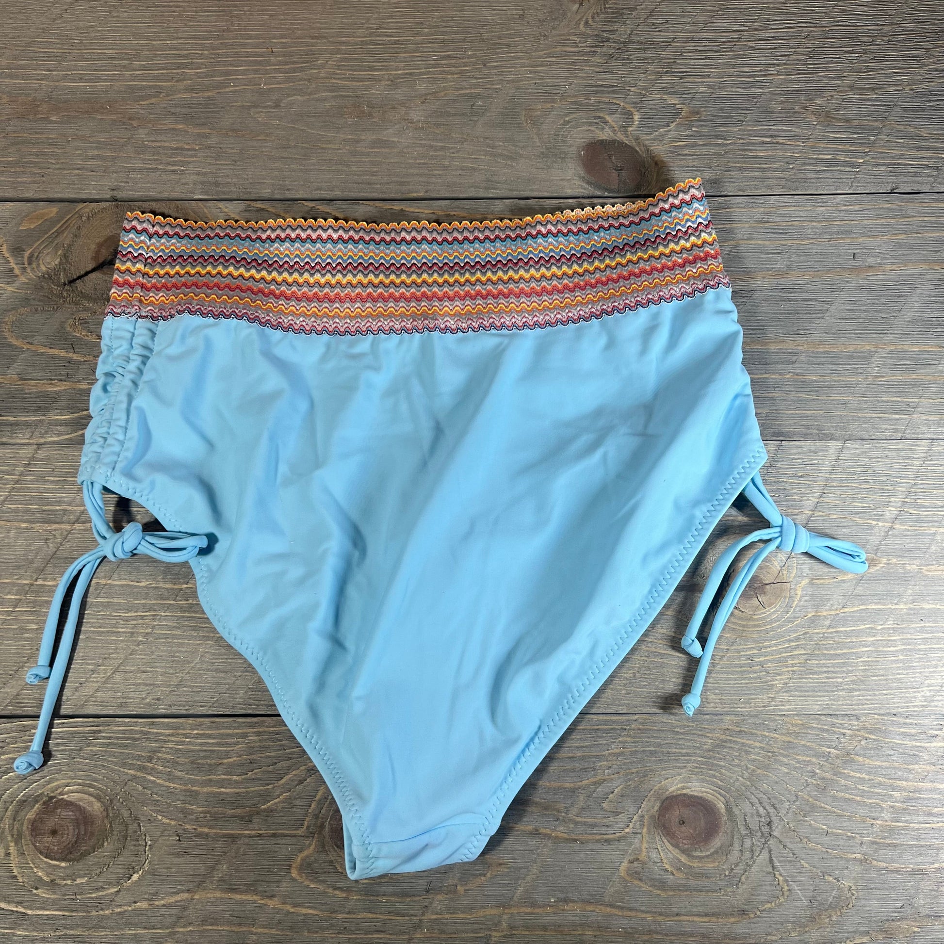 Light blue high waisted bikini bottom with colorful zigzag waistband and side ties on wooden background. Size M.
