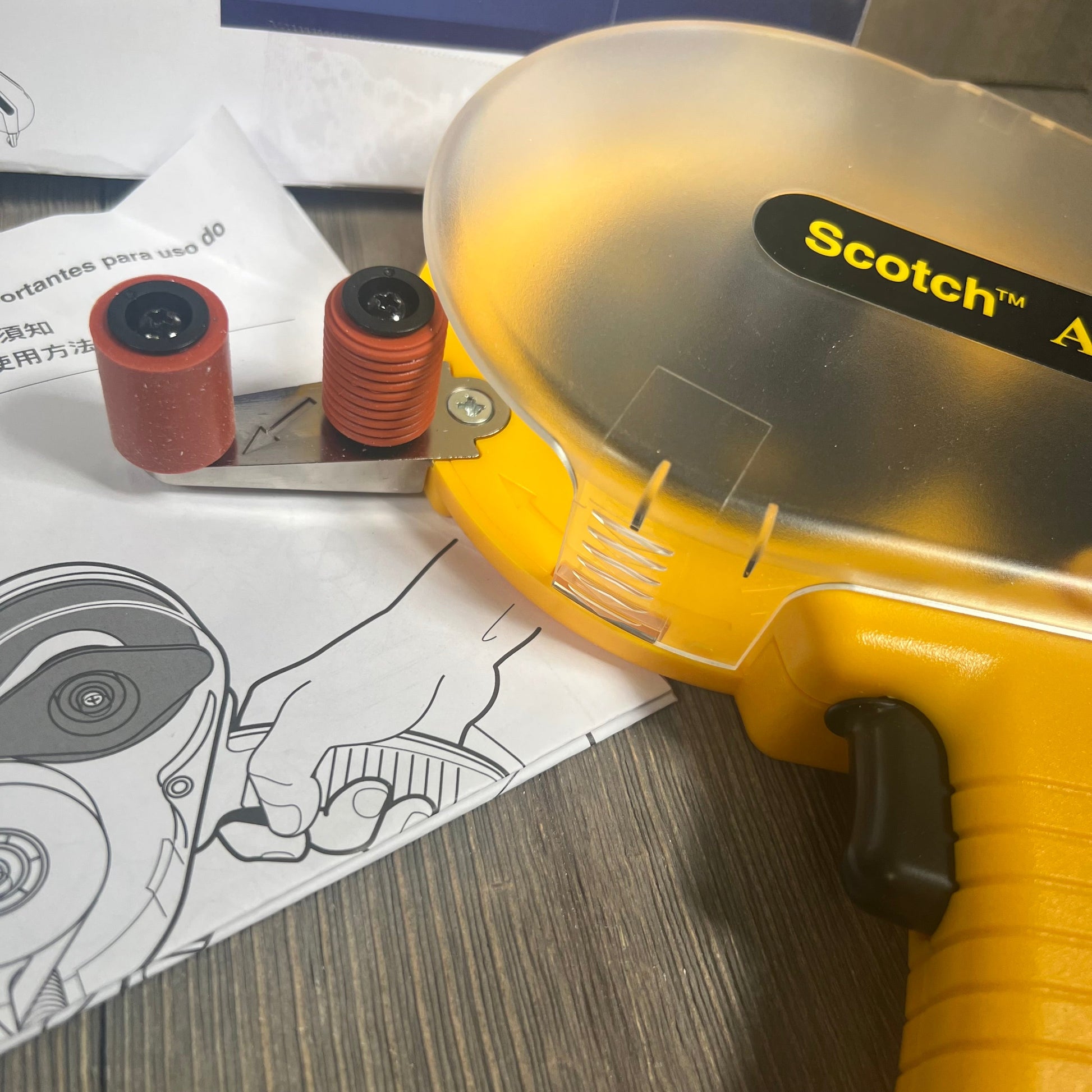 Close-up of the 3M Scotch ATG 700 Adhesive Applicator in yellow, showing rollers and instructions on a wooden surface.