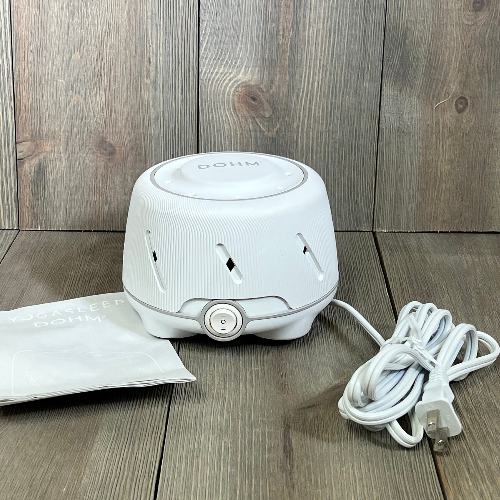 White Yogasleep Dohm noise machine, fan-based, with power cord on wooden surface, ideal for sleep and meditation.