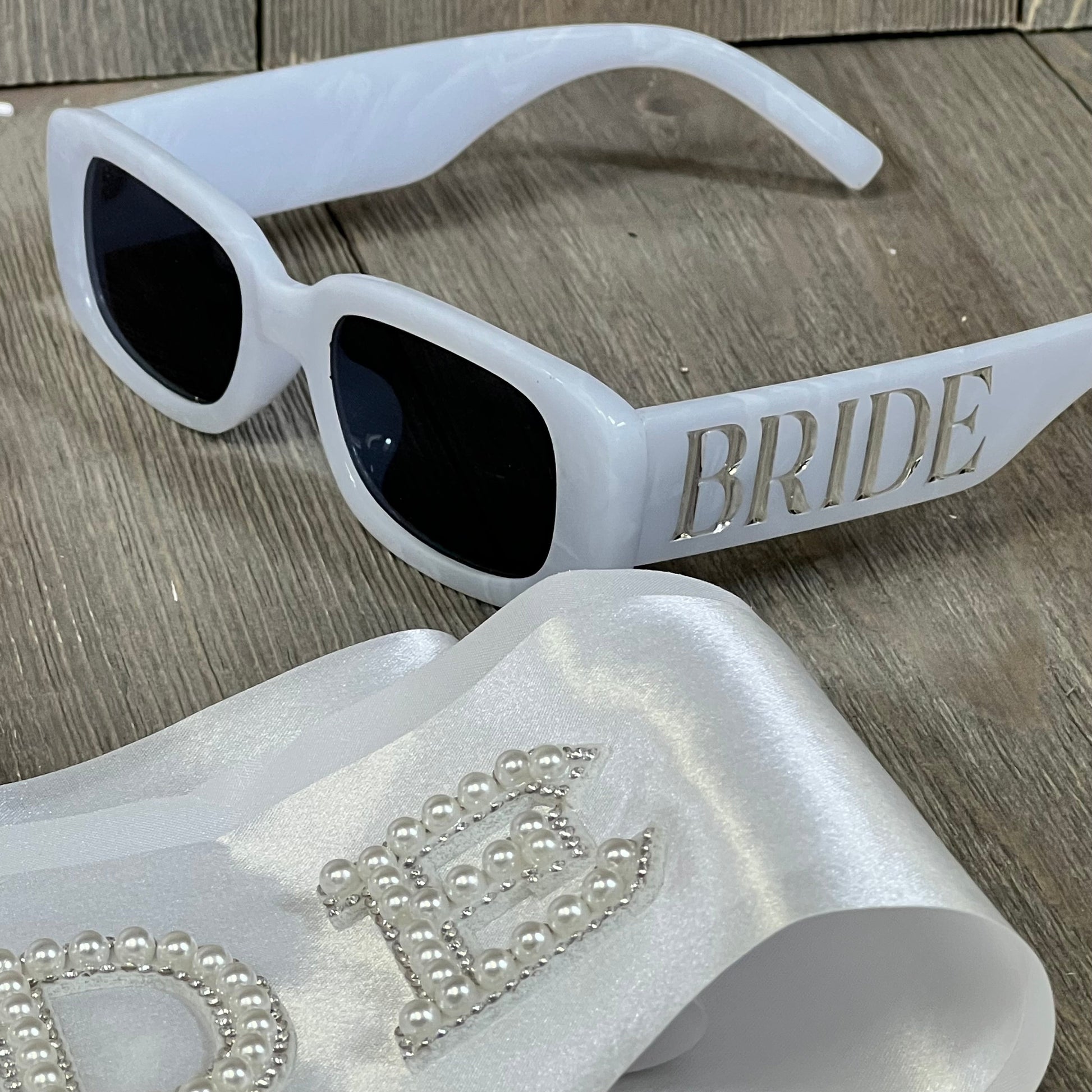 White bridal sunglasses with "BRIDE" lettering, paired with a pearl-embellished satin sash, perfect for wedding celebrations.