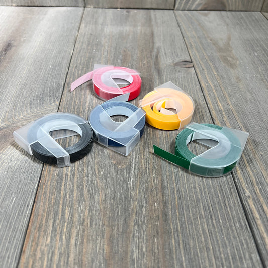 Five rolls of embossing label tape in black, blue, red, yellow, and green on a wooden surface.