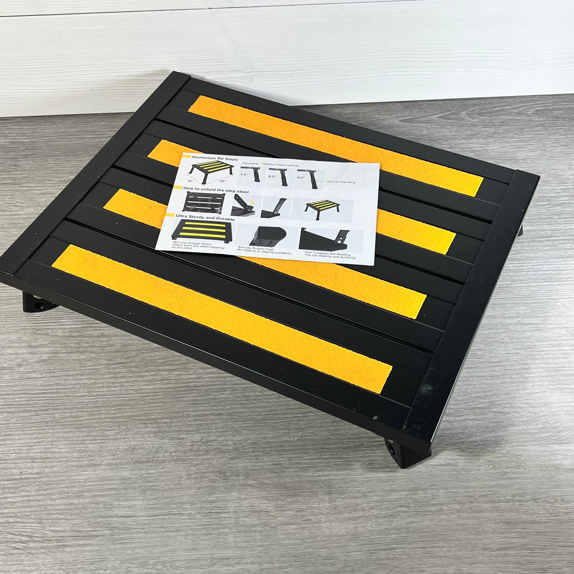 Adjustable black aluminum RV step with non-slip yellow grips, includes instructional leaflet on a light wood floor.