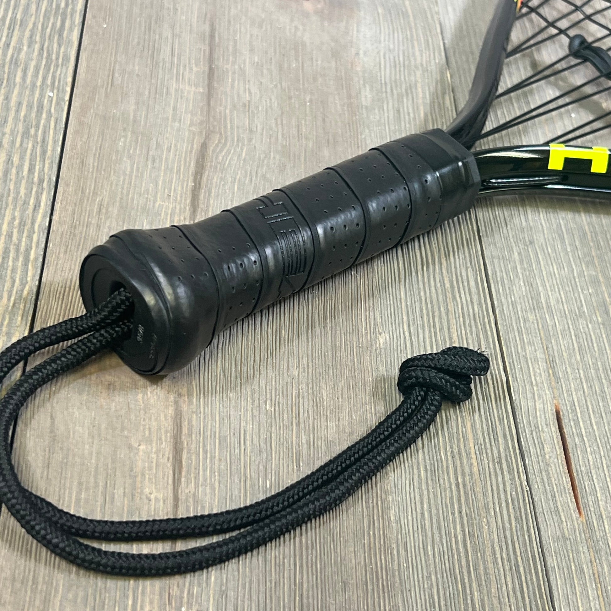 Close-up of 1047180423-HEAD CPS Heat Racquetball Racquet handle, featuring a black grip and wrist strap on wood surface.