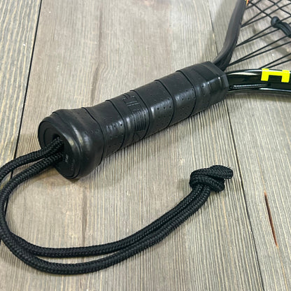 Close-up of 1047180423-HEAD CPS Heat Racquetball Racquet handle, featuring a black grip and wrist strap on wood surface.