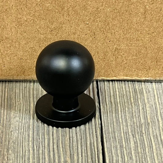 Matte black 1-inch kitchen cabinet knob on textured wood background, ideal for modern dresser drawers.