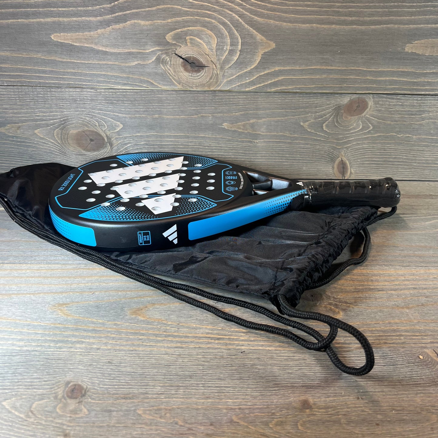 Blue Adidas Rx 2000 Light padel racket on a wooden surface with a black carrying bag.