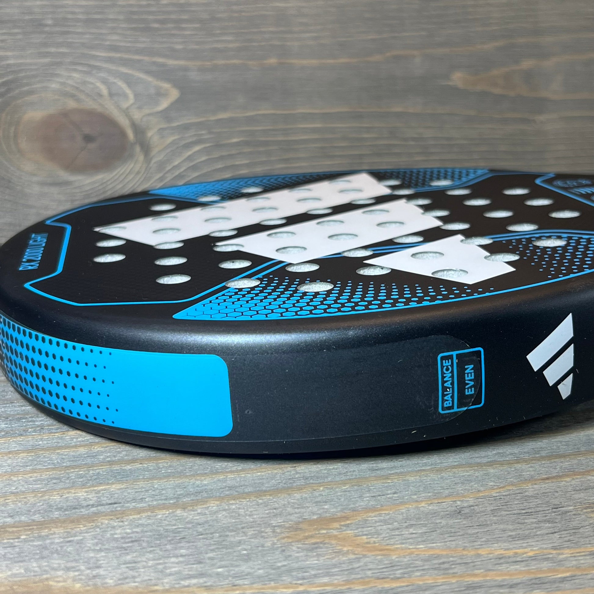 Blue Adidas Rx 2000 Light padel racket on wooden surface, showcasing perforated design and even balance label.