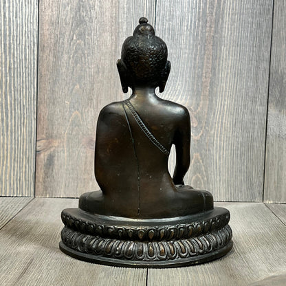 Bronze Buddha statue viewed from the back, showcasing intricate robe details and serene craftsmanship against a wooden backdrop.