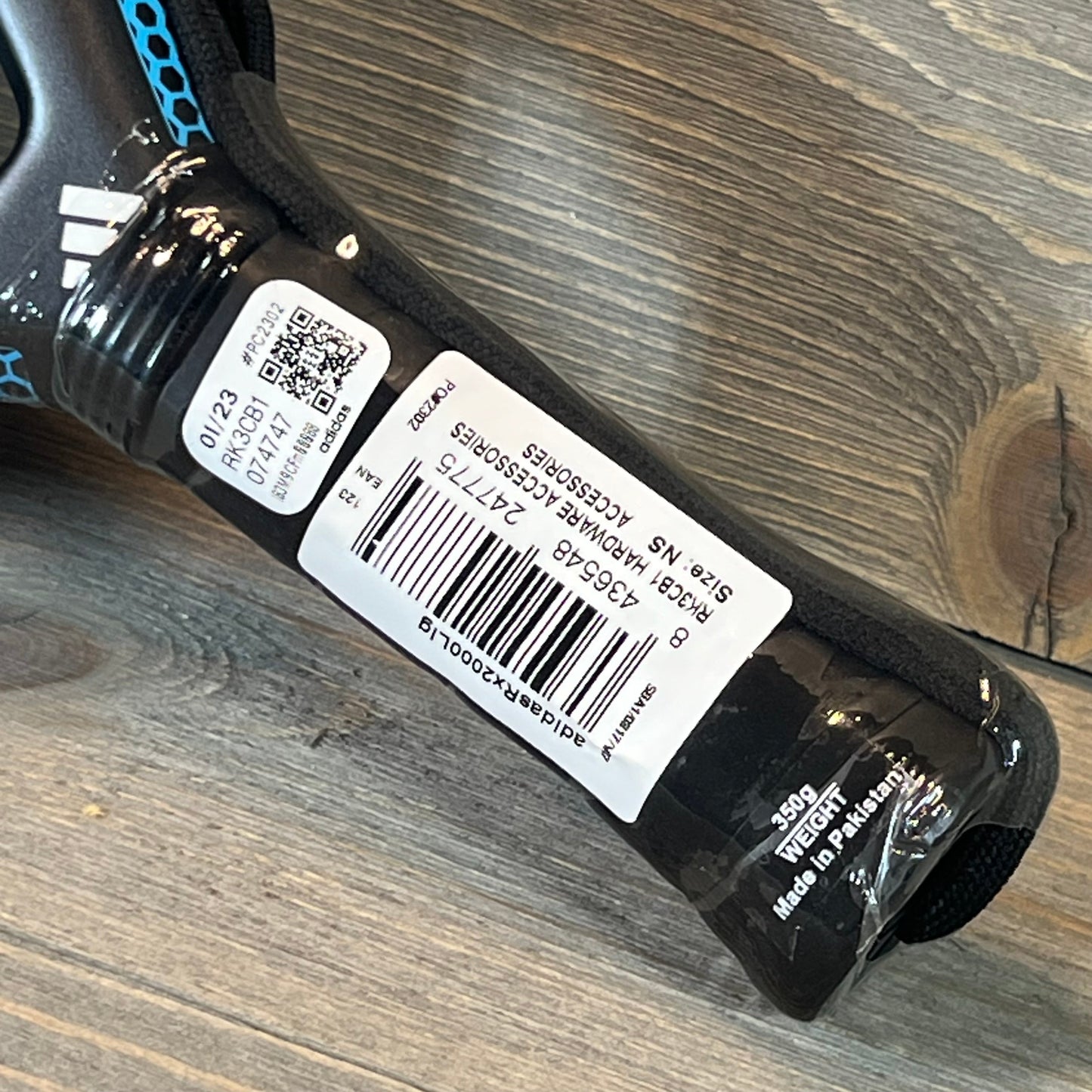 Close-up of Adidas Rx 2000 Light Padel Racket handle showing barcode and product details on a wooden surface.