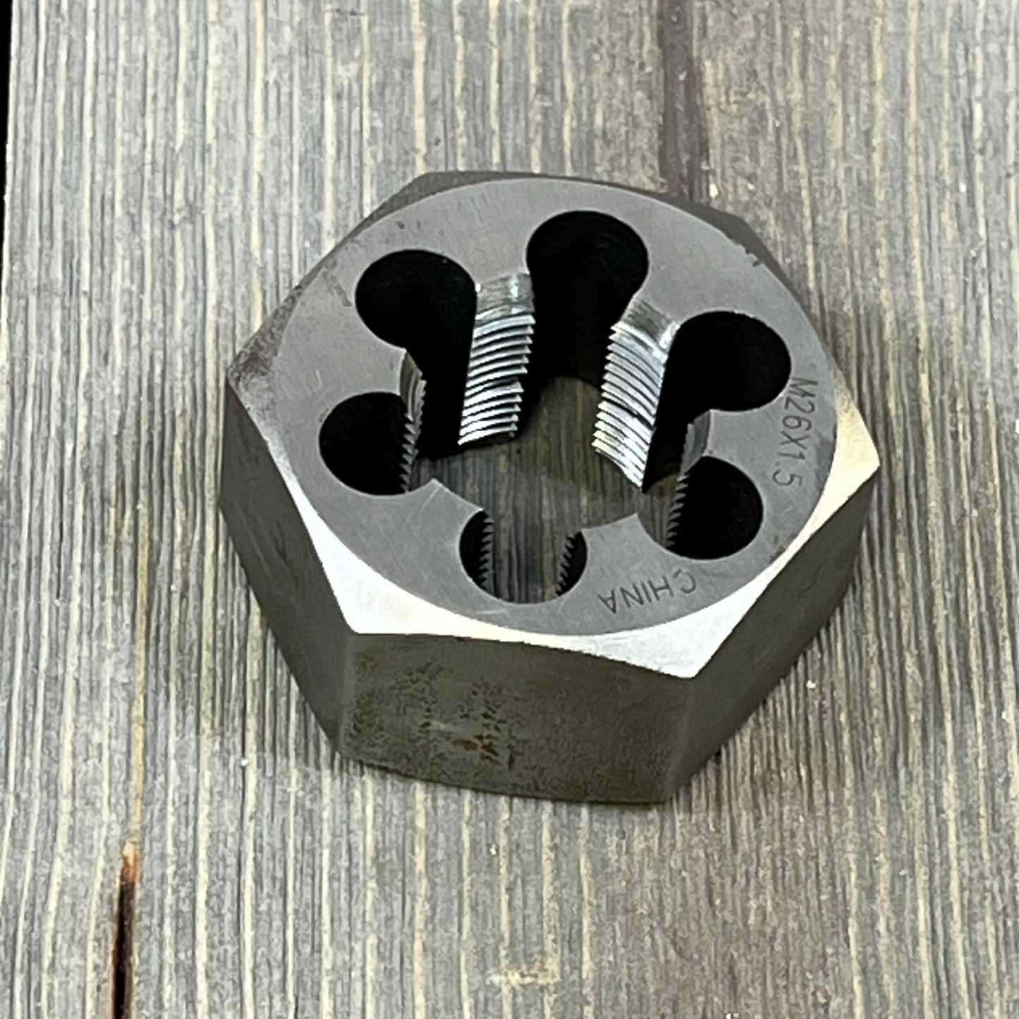 Hexagonal carbon steel rethreading die with sharp threading, size M26 x 1.5, resting on a wooden surface.