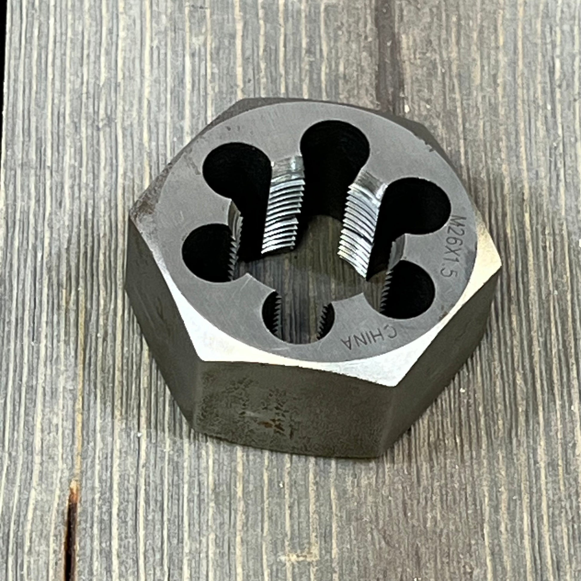 Hexagonal carbon steel rethreading die with sharp threading, size M26 x 1.5, resting on a wooden surface.