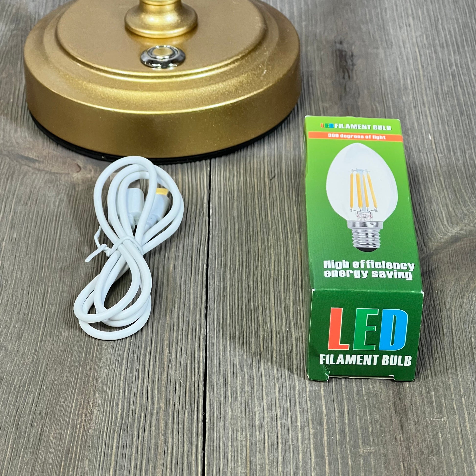 Gold rechargeable table lamp base with touch control, LED E12 bulb packaging, and charging cord on wooden surface.