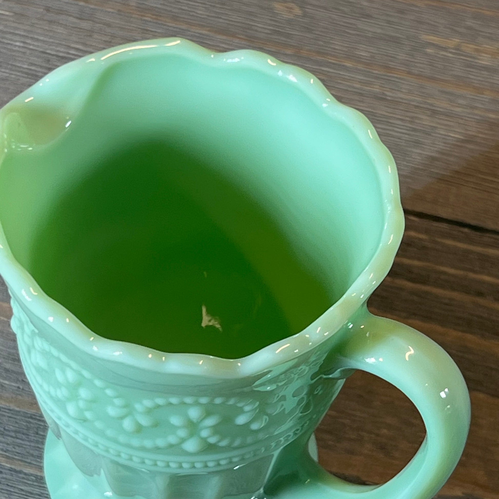 Mint green creamer with intricate floral pattern, scalloped edge, and elegant handle on rustic wooden surface.