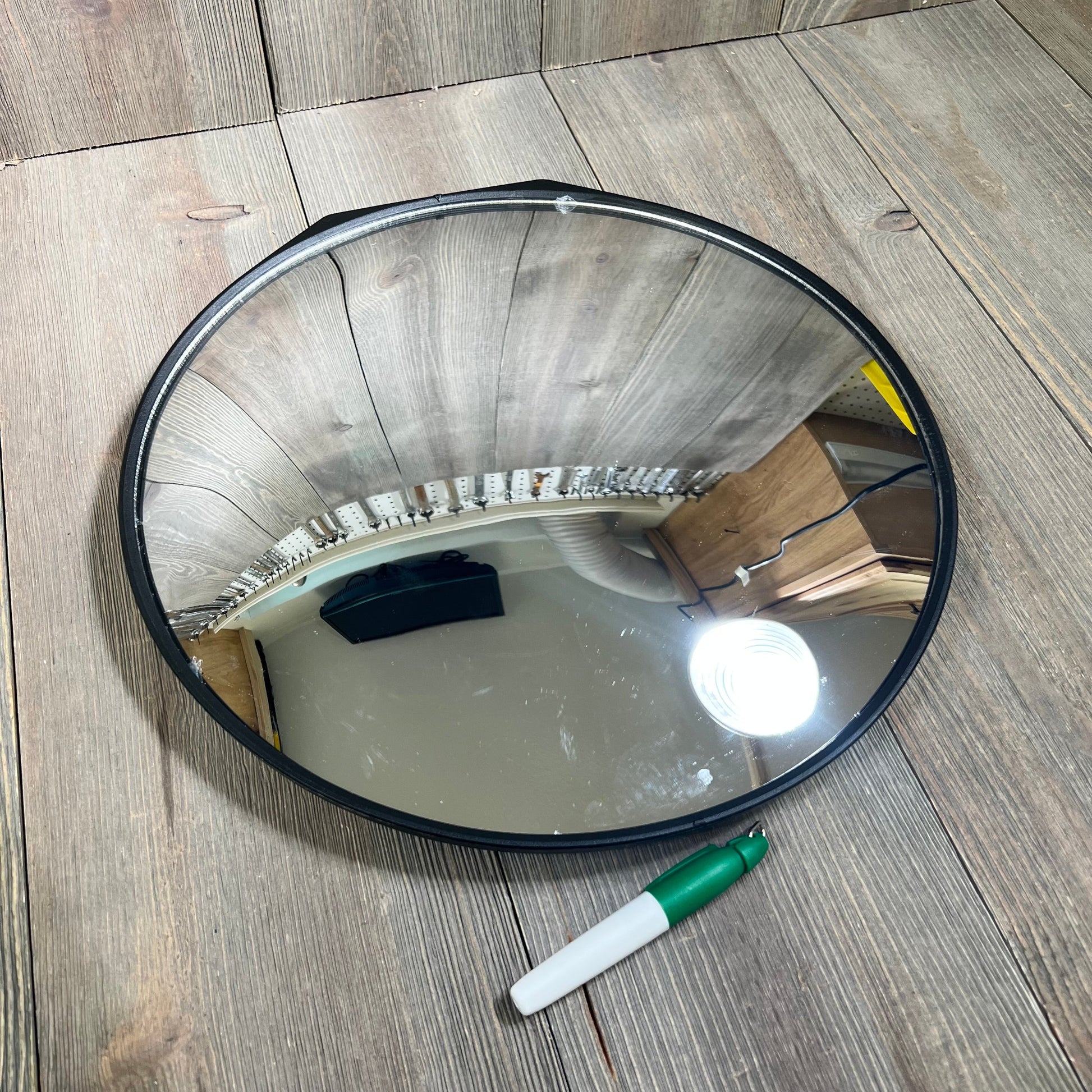Round golf swing training mirror on a wooden surface, reflecting light and nearby objects, with a green marker beside it.