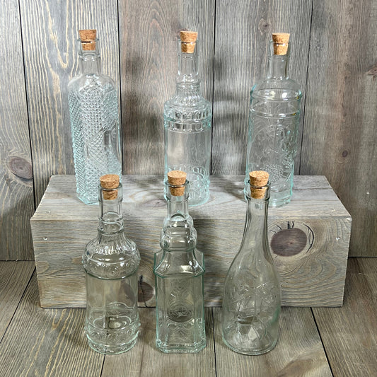 Assorted clear glass bottles with corks on wooden surface, 6 pack, 16oz, 2.5x9in, ideal for decor or storage.