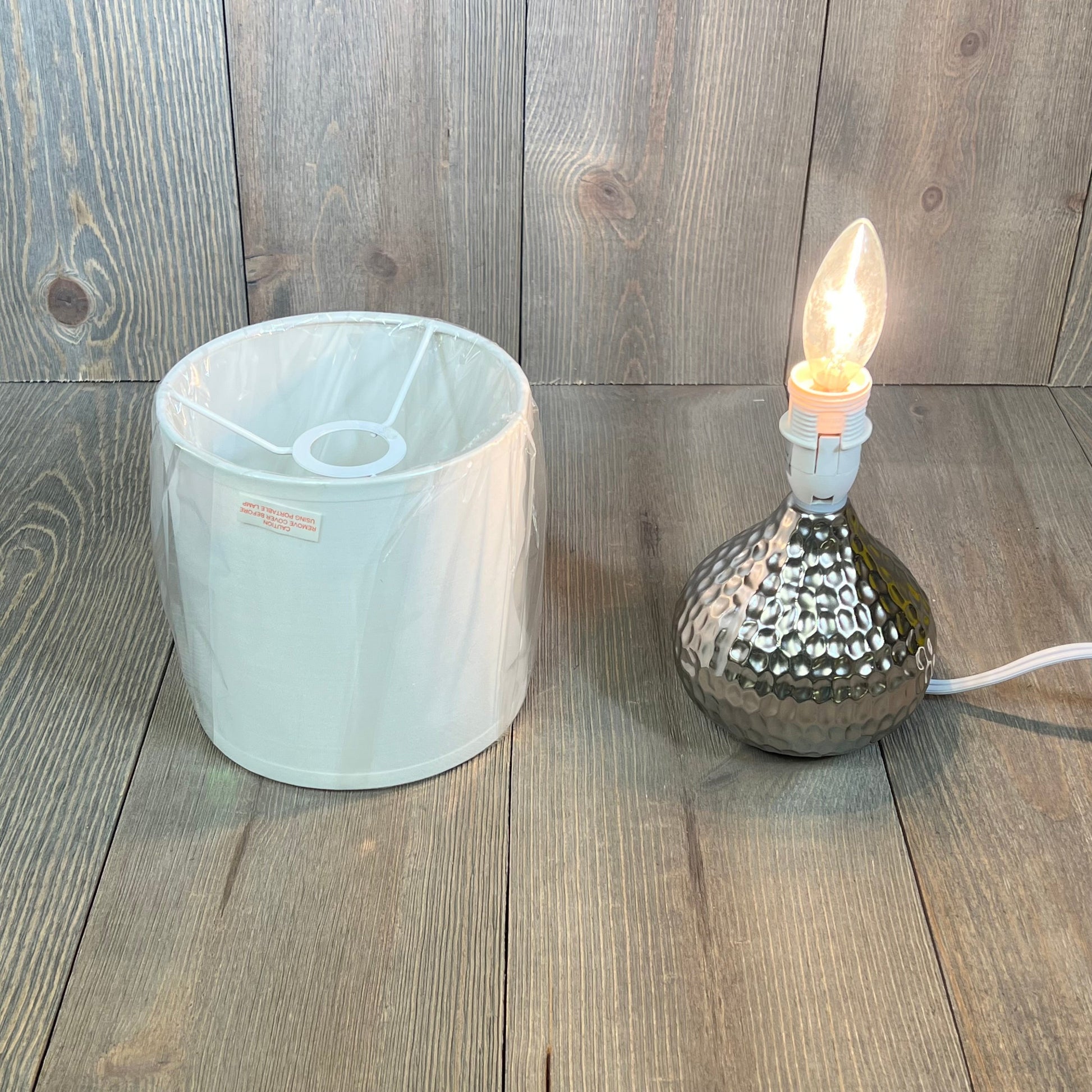 Mini hammered texture silver table lamp with exposed bulb and detached white shade on wooden surface.