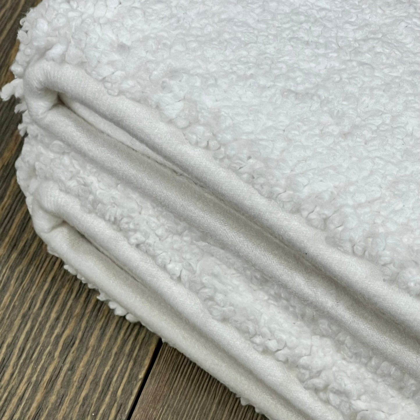 Soft, white DEXI bathroom rug folded on wooden floor, showcasing plush microfiber texture and quality craftsmanship.