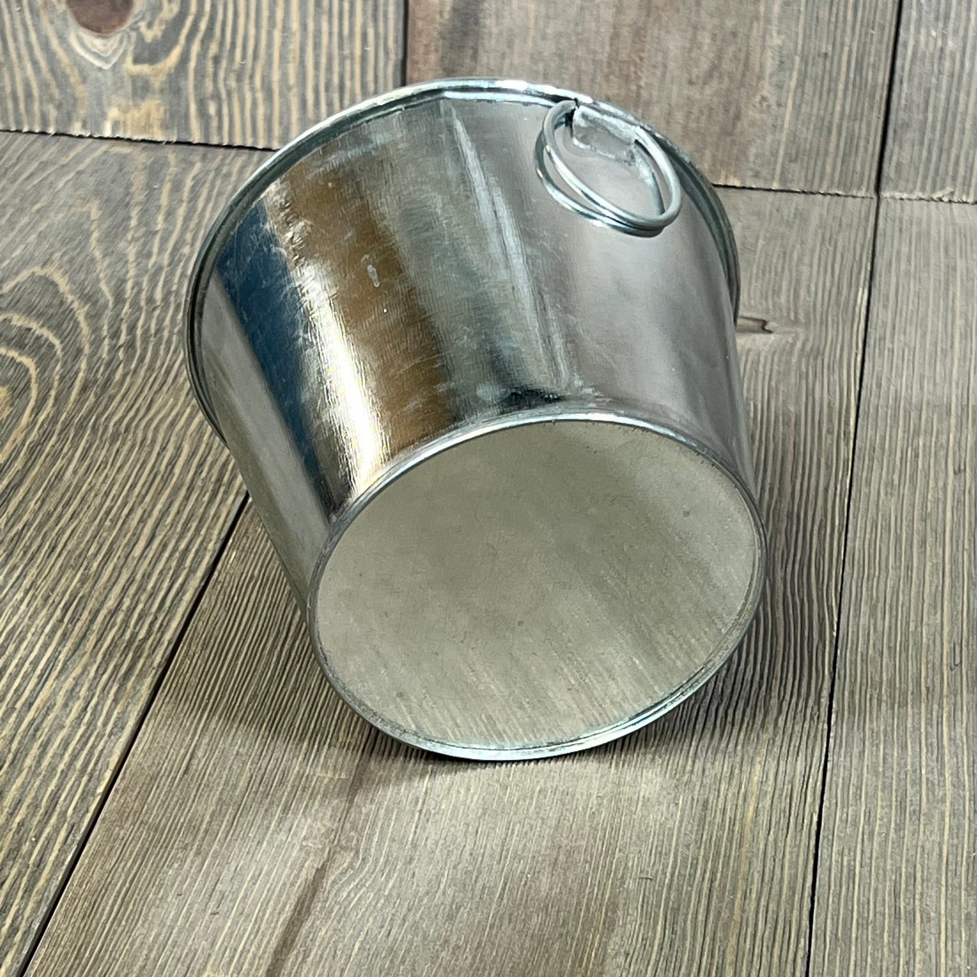 Mini galvanized bucket with handle on wooden surface, ideal for rustic decor or gardening use. Size: 4.5 x 3.5 inches.